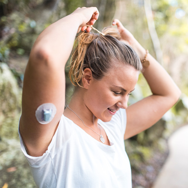 Woman wearing a continuous glucose monitor on her upper arm while tying her hair back outdoors, showing active diabetes management.