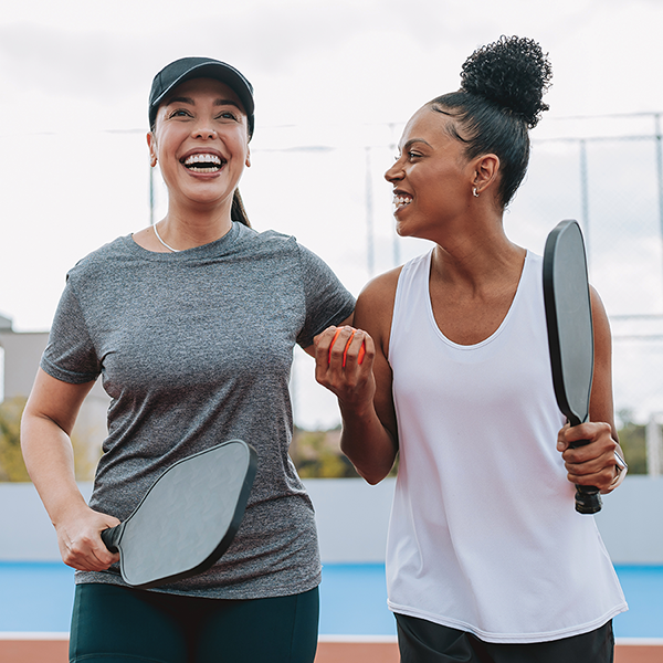Two healthy women smiling and playing pickleball outdoors, enjoying an active lifestyle.