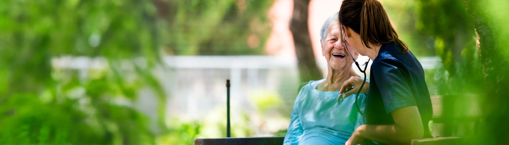 A nurse and an older adult share a joyful moment on a bench outside, surrounded by greenery and a sense of compassionate care.