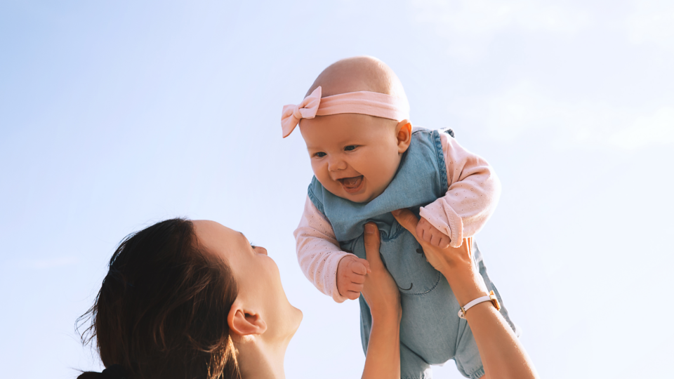 Smiling baby lifted in the air by mom, illustrating wrist strain that can occur from repetitive lifting after childbirth.