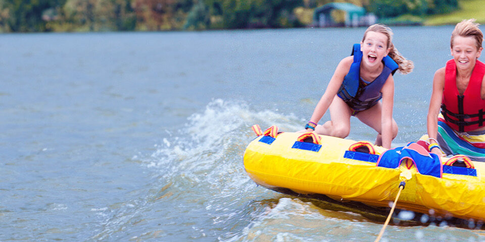 Two smiling kids wearing life jackets enjoying a safe summer tubing adventure on a lake.