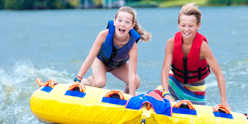 Two smiling kids wearing life jackets enjoying a safe summer tubing adventure on a lake.