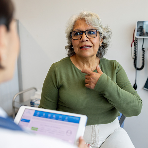 Older woman at a doctor’s appointment pointing to her throat, describing GERD and acid reflux symptoms.
