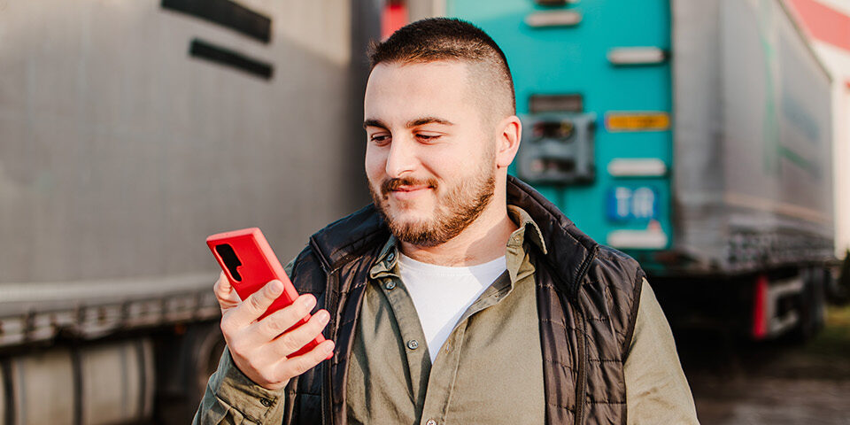 Man using phone to schedule a DOT physical offered at CRMC, holding clipboard while standing near trucks.