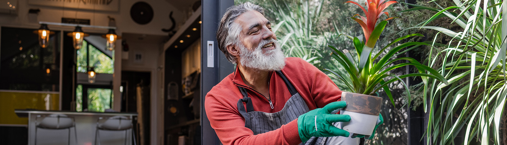 Older man smiling while gardening and holding a potted plant, representing a healthy back, strength, and active lifestyle.