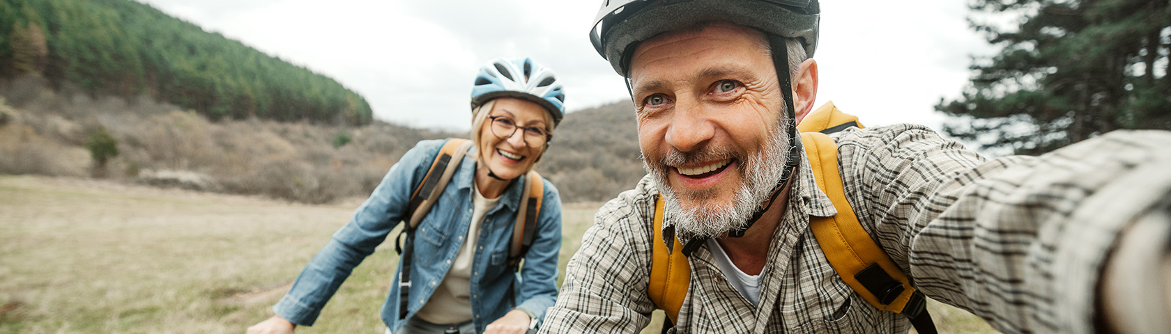 Smiling older couple wearing bike helmets and backpacks on a trail, representing back health, posture, and active outdoor living.