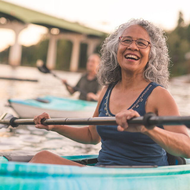 Smiling woman kayaking on the water, representing a healthy back, upper body strength, and active, pain-free living.