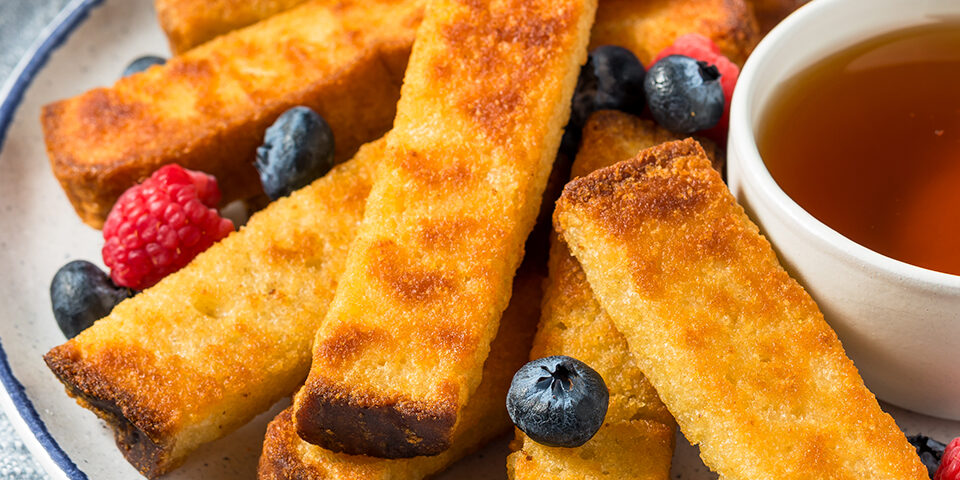 Stack of golden keto French toast sticks drizzled with sugar-free syrup and topped with fresh berries, styled on a rustic breakfast plate.