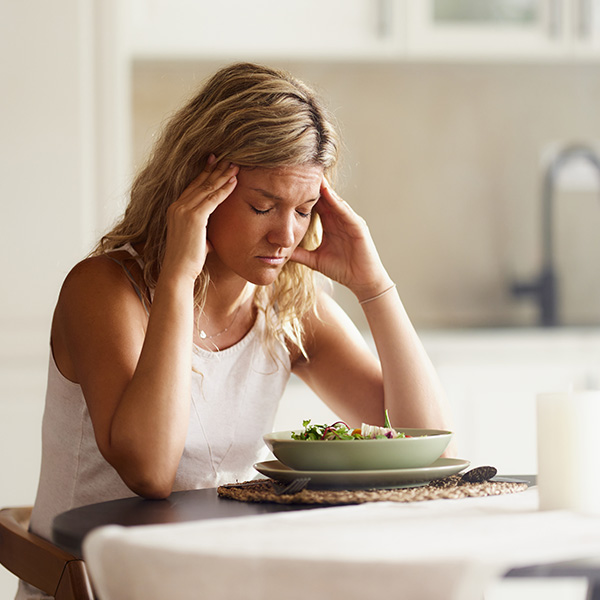 Woman holding her head in discomfort while sitting in front of a meal, showing symptoms of gastroparesis.