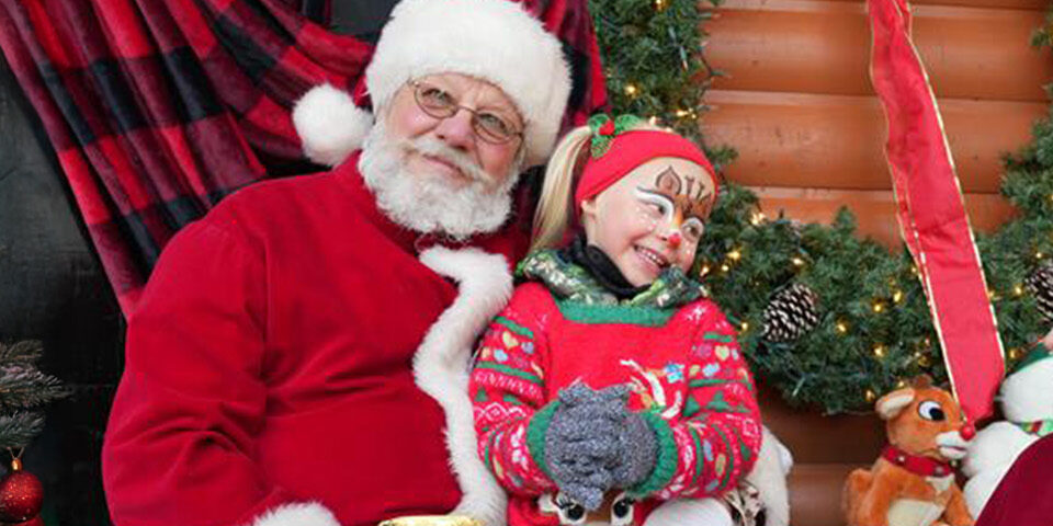 A smiling child with face paint sits with Santa and Mrs. Claus in front of a holiday wreath during CRMC’s Snow Many Smiles event in Crosby.