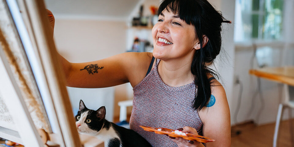 Person painting at an easel with a cat nearby, representing creative hobbies and self-care supported by CRMC Behavioral Health for a healthy mindset.
